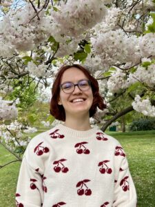 Kaja Grimme, eine junge weiße Frau mit kinnlangen welligen dunkelroten Haaren, steht unter den blühenden Zweigen eines Kirschbaums. Sie trägt einen weißen Pullover mit roten Kirschen darauf und lacht.