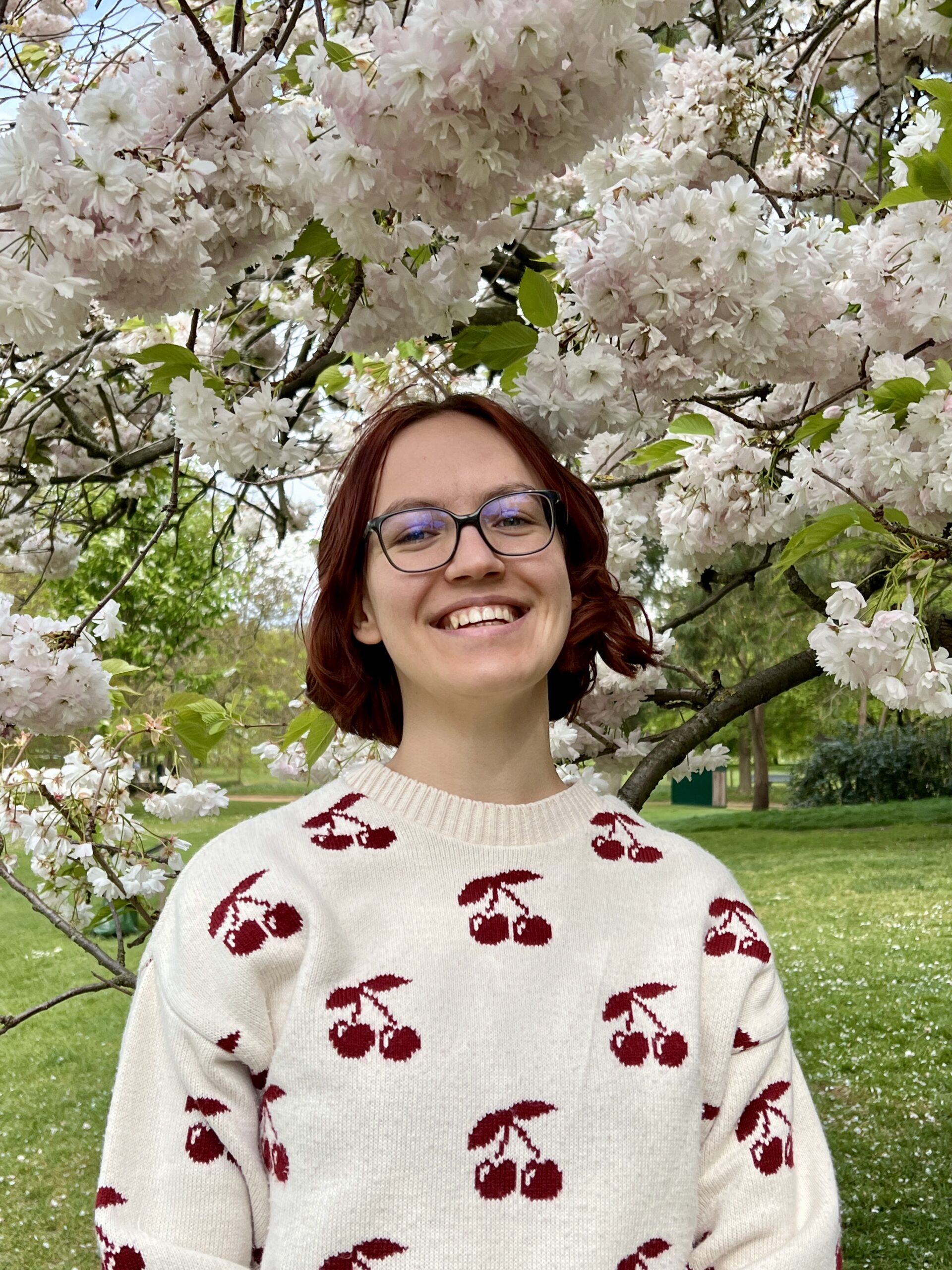 Kaja Grimme, eine junge weiße Frau mit kinnlangen welligen dunkelroten Haaren, steht unter den blühenden Zweigen eines Kirschbaums. Sie trägt einen weißen Pullover mit roten Kirschen darauf und lacht.
